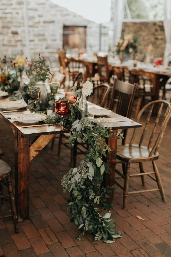 A rustic dining table decorated with green foliage and white flowers, set for a celebration.