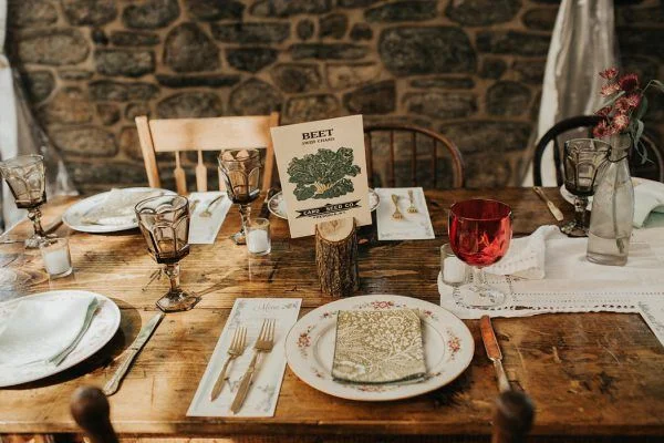 A rustic dining table set with vintage plates, glasses, and utensils, decorated with a card labeled 'Beef' and a small tree log for a centerpiece, with a stone wall background.