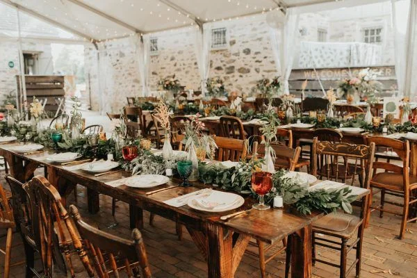 Long wooden dining tables with white plates, glassware, and festive table runners decorated with greenery inside a white tent with brick flooring.