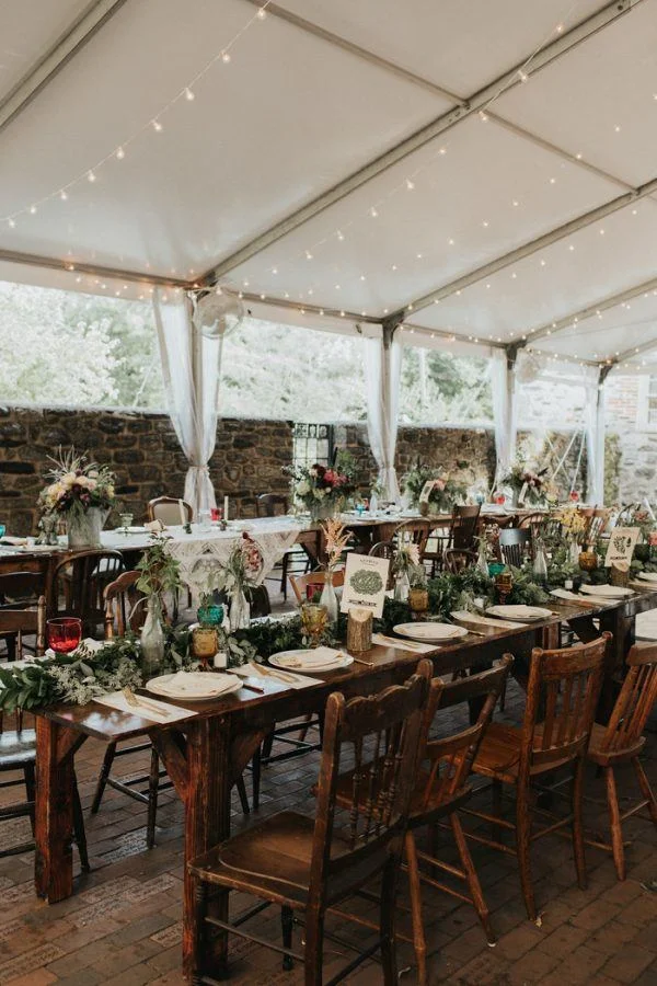 A decorated outdoor event space under a white tent with wooden tables, chairs, floral centerpieces, and string lights.
