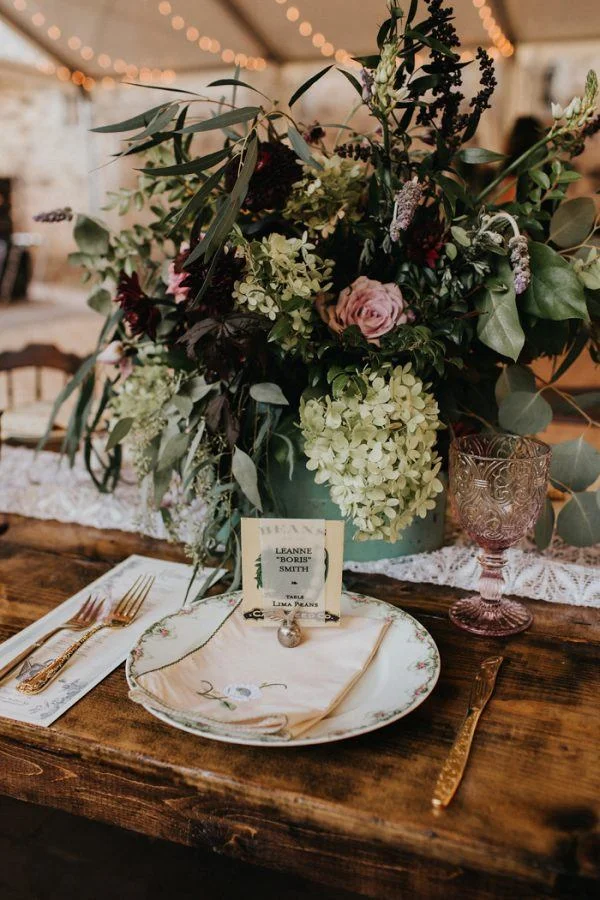 A rustic table setting with a centerpiece of white and pink flowers, greenery, and eucalyptus in a blue vase, with a white lace runner and gold utensils.