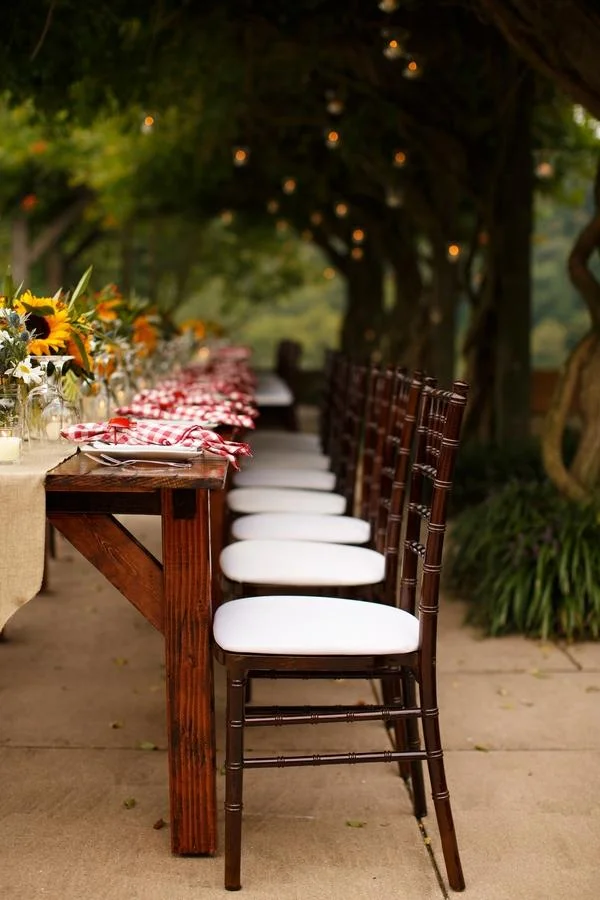 Outdoor dining table with white cushioned chairs, rustic wooden table with red checkered napkins, sunflower centerpiece, under string lights in a garden setting.