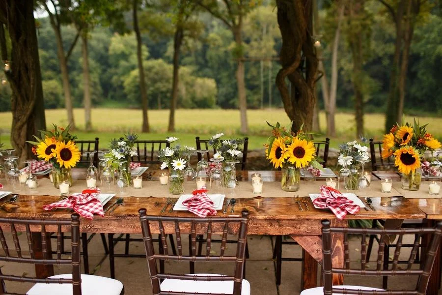 Long wooden table decorated with sunflower and daisy floral arrangements, red and white checkered napkins, and small candles, set outdoors in a park with green grass and trees in the background.