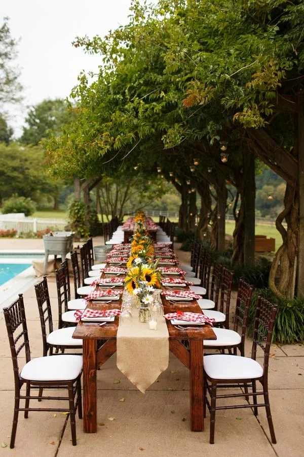 A long outdoor dining table set for a gathering, decorated with sunflower centerpieces and red checkered napkins, positioned under a canopy of lush green trees near a swimming pool.