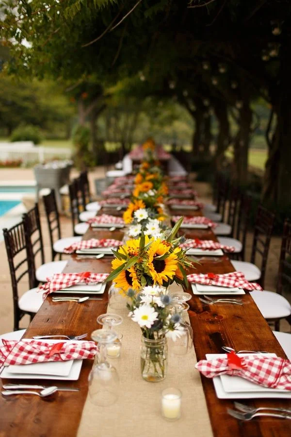 A long outdoor dining table decorated for a gathering, with sunflower and daisy floral centerpieces, white plates, silverware, red and white checkered napkins, and small glass jars with candles, set under a canopy of trees.