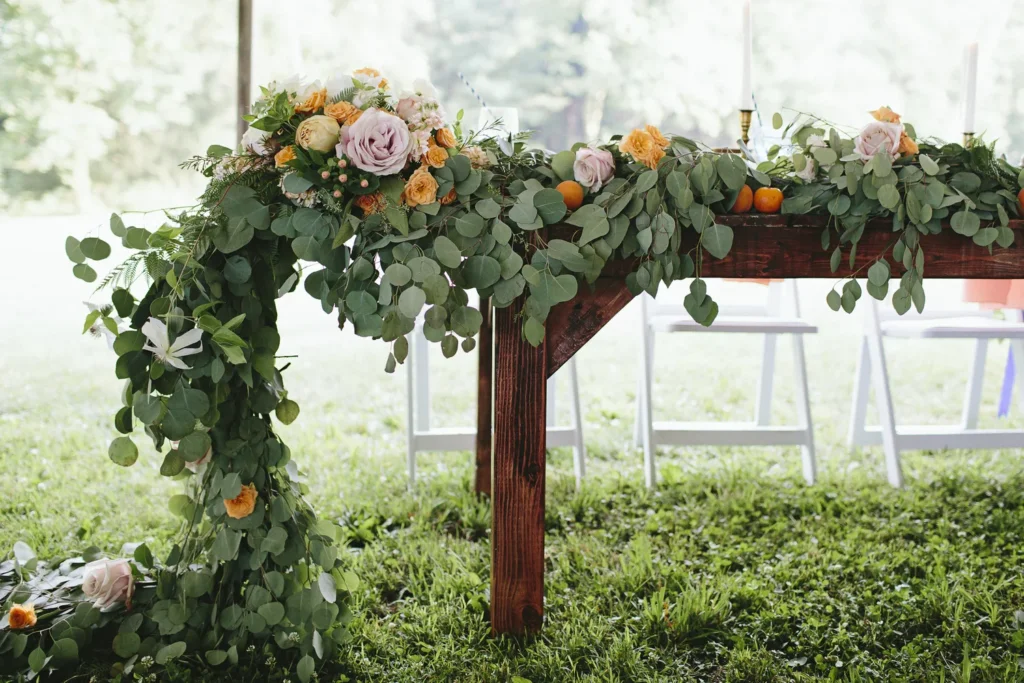 A floral arrangement with pink and orange flowers and green foliage on a wooden table, outdoors with chairs and candles in the background.