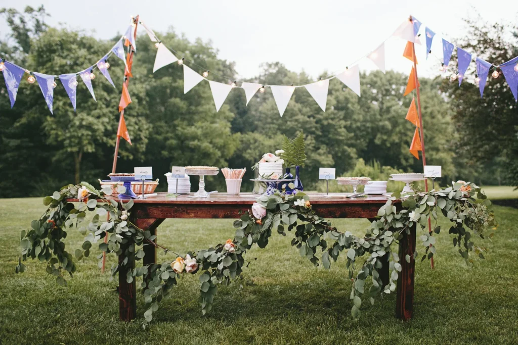 Outdoor dessert table decorated with green foliage and flowers, set up for a celebration with bunting flags in purple, white, and orange.