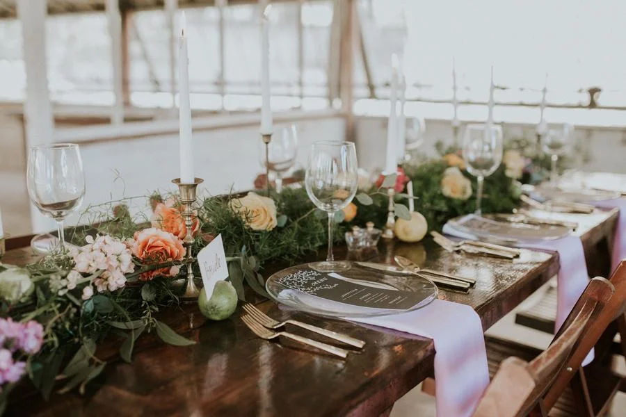 Elegant dining table set with candles, wine glasses, floral centerpiece, plates, and silverware in a bright indoor setting.