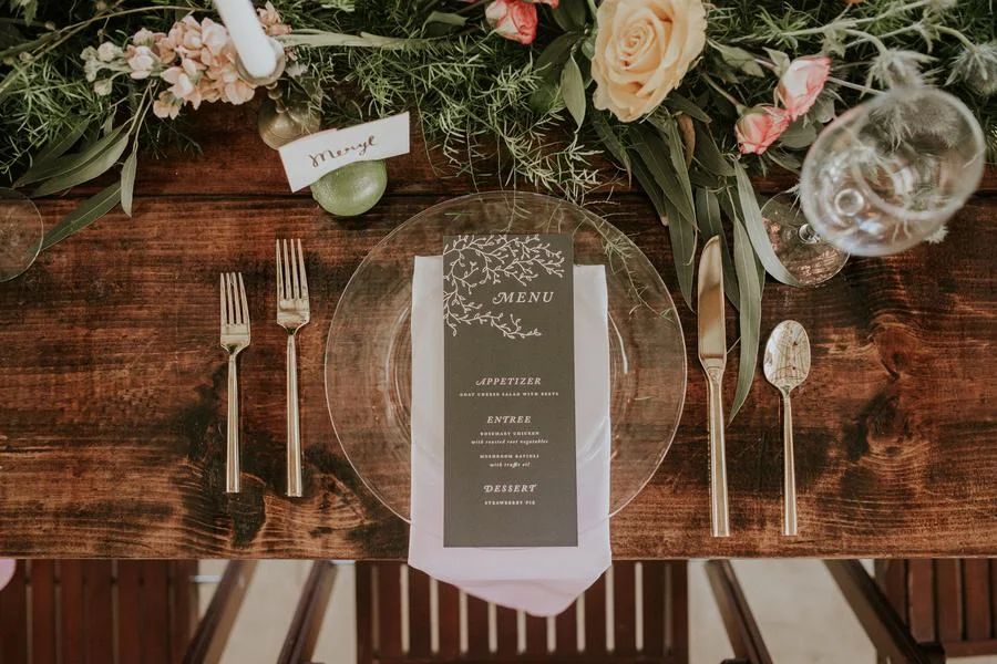 Elegant table setting with a clear glass plate, white napkin, and a menu card in the center, with a floral centerpiece of roses and greenery.
