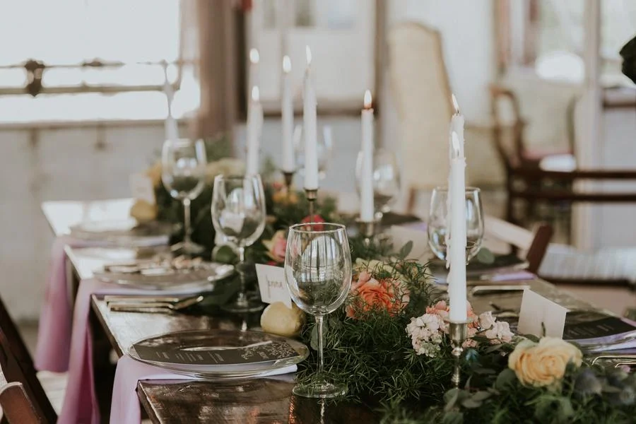 A long dining table decorated with pink and white flowers, greenery, and tall white candles in silver candle holders, set for a formal event or celebration.
