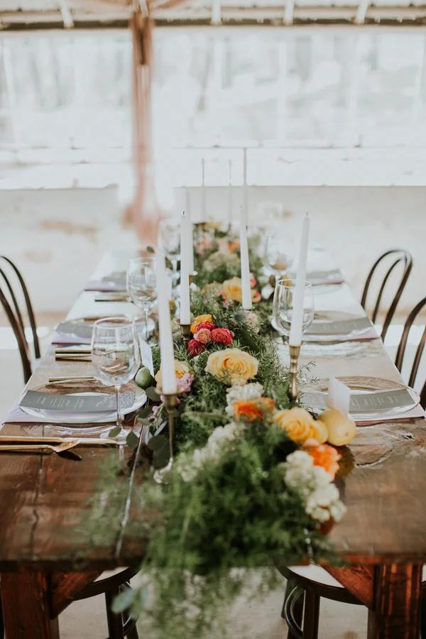 A long dining table with a floral centerpiece, candles, wine glasses, and neatly arranged place settings, set for a formal event in a bright, airy room.