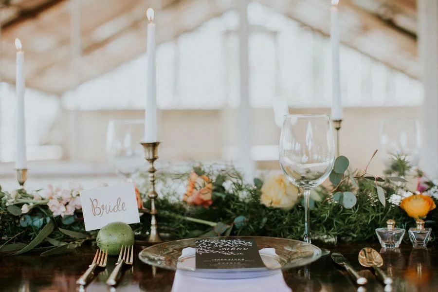Elegant wedding table setting with a 'Bride' sign, tall white candles, wine glasses, greenery, and flowers.