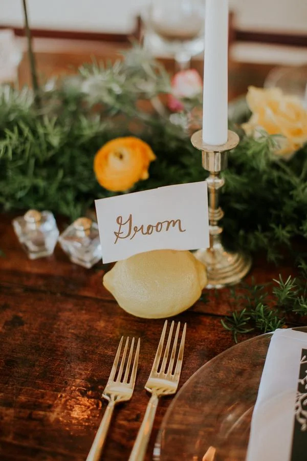 A table setting with a lemon, a white candle in a silver holder, a small card with the word 'Groom' written in cursive, surrounded by greenery and flowers.
