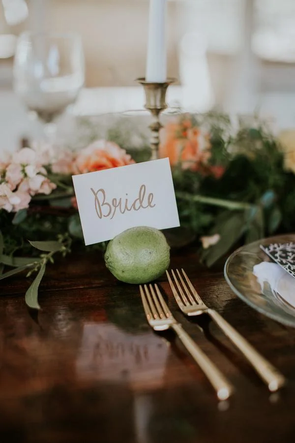 Wedding reception table setting with a lime holding a 'Bride' sign, gold forks, a floral centerpiece, and candle.