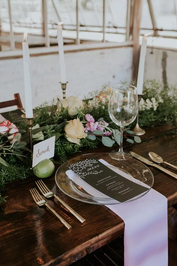 A formal dining table decorated with a greenery and flower centerpiece, white candles, a table number card, a printed menu on a plate, a white table runner, wine glass, and gold flatware.