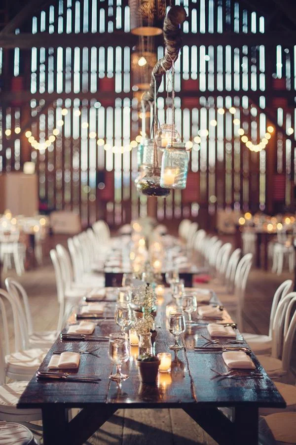 Long wooden banquet table set with wine glasses, napkins, and candles in a rustic event space with string lights and hanging jars.
