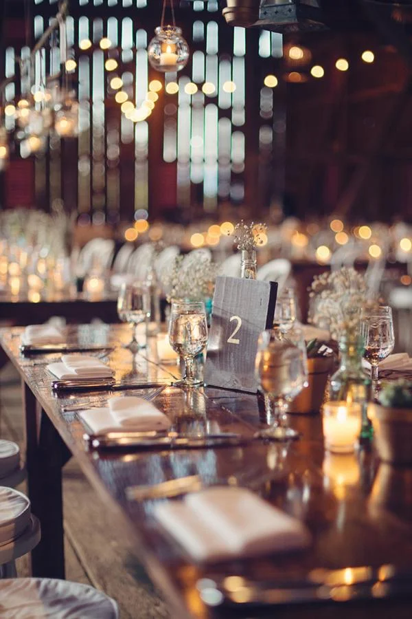 A decorated dining table set for an event in a rustic venue with string lights and candles, featuring glasses, napkins, and a table number card.