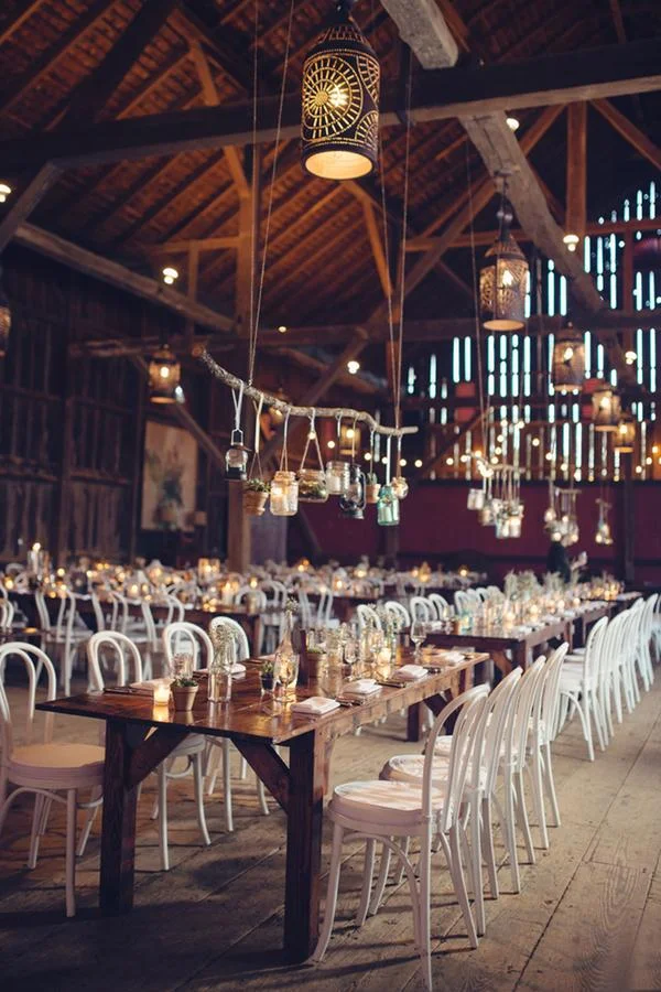 Wedding or event reception in a rustic barn with wooden tables and white chairs, decorated with candles, jars, and hanging lights.