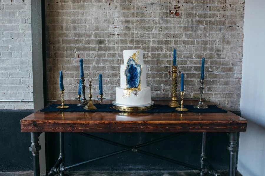 Cake with blue geode design on a wooden table, surrounded by gold and brass candlesticks with blue candles, against a brick wall.
