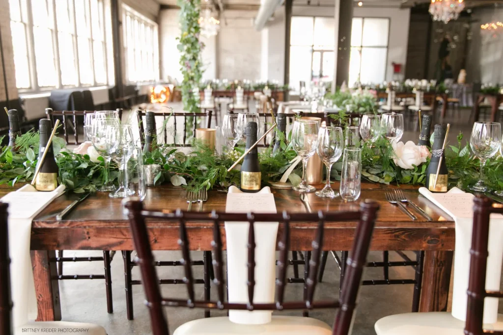 Wedding reception table decorated with candles, greenery, wine glasses, champagne bottles, and place cards in a rustic, indoor venue with natural light and hanging lights.