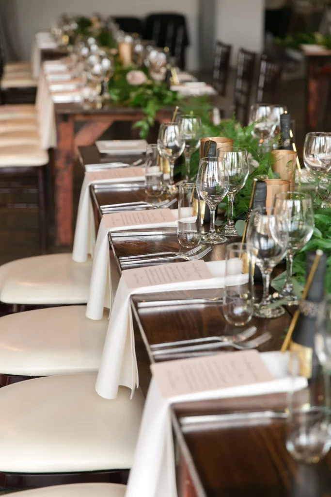 A wedding reception table set with white napkins, wine glasses, and menus, decorated with green foliage and pink flowers.