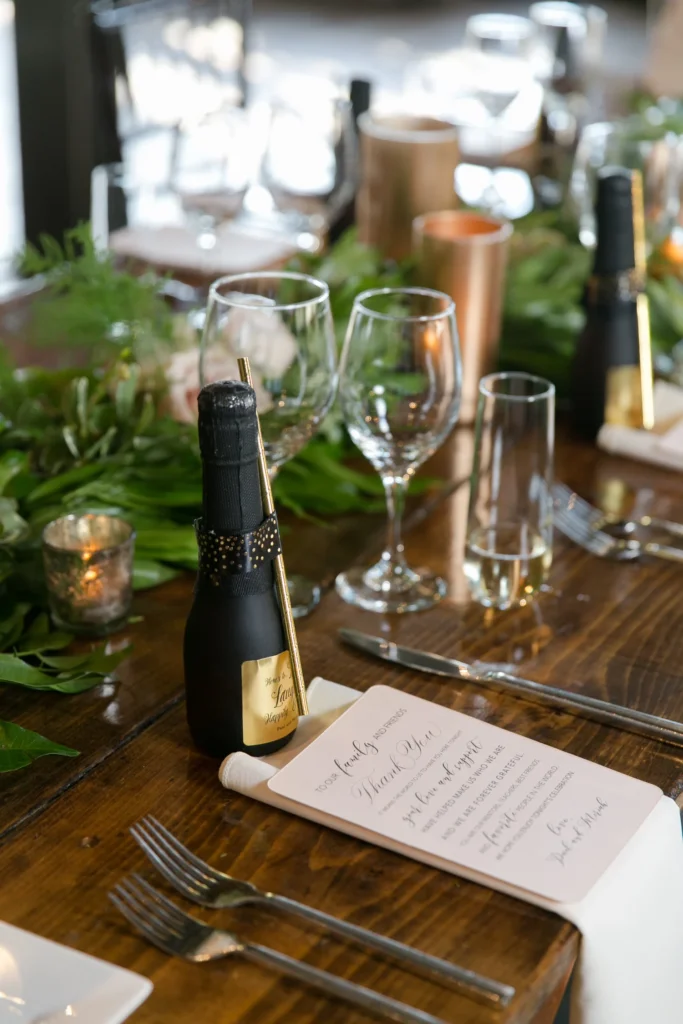 A decorated dining table set for a celebration with empty wine glasses, candles, green foliage, a small bottle resembling champagne, and a printed thank you note.
