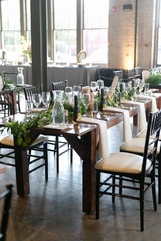 Wedding reception table with floral centerpiece, wine glasses, water bottles, and champagne bottles in a rustic venue with large windows.