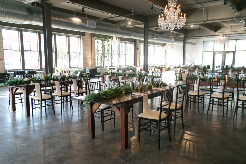 An elegantly decorated event space with wooden tables surrounded by black and white chairs, lush greenery centerpieces, glassware, and candles, illuminated by natural light from large windows.