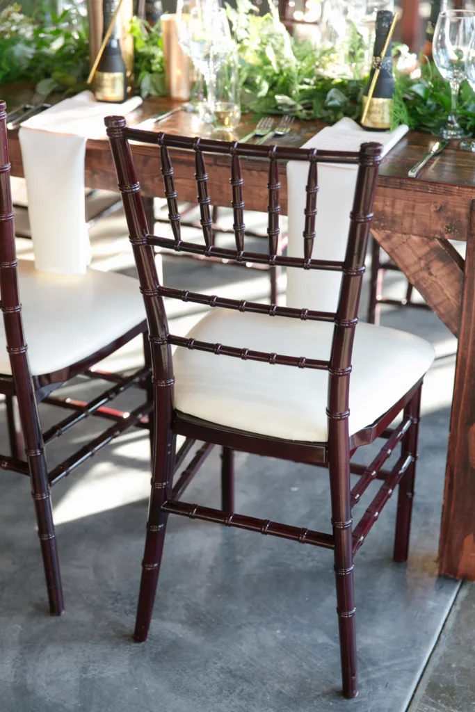 Close-up of a dark wooden chair with a white cushion, part of a dining table set with white cloth napkins, glassware, and greenery in the background at a restaurant or event venue.