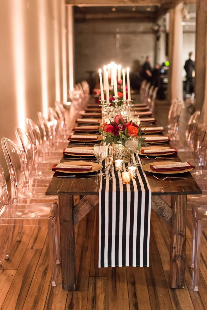 Long wooden dining table decorated with floral centerpieces, candles, black and white striped table runner, and place settings with glasses and napkins, in a warmly lit rustic venue.