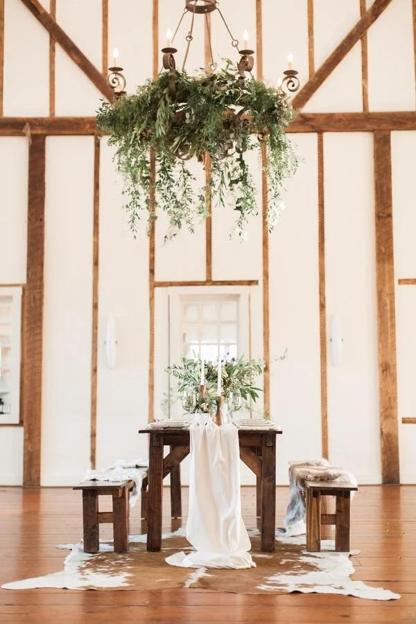 Rustic dining room with wooden table and benches, a cowhide rug, candles, and hanging greenery chandelier.