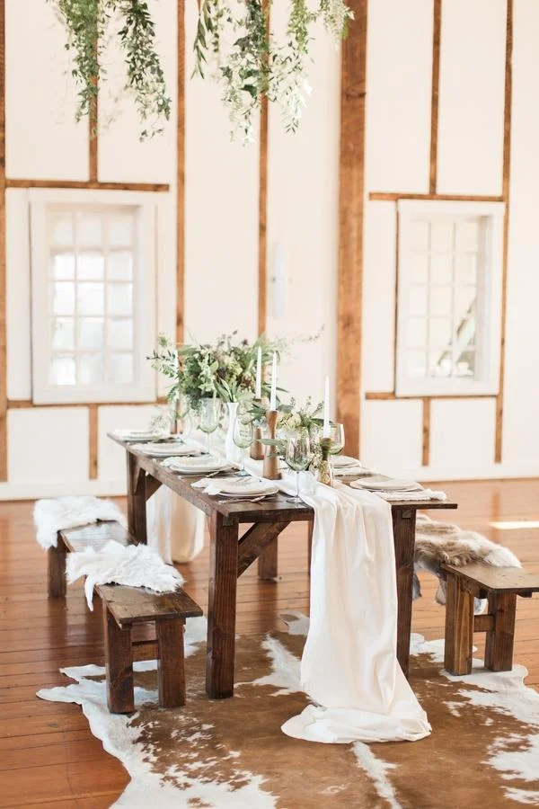 Rustic wooden table set for a formal event with white tablecloth and greenery centerpiece, surrounded by matching benches with sheepskin throws, in a bright room with white walls and wood accents.