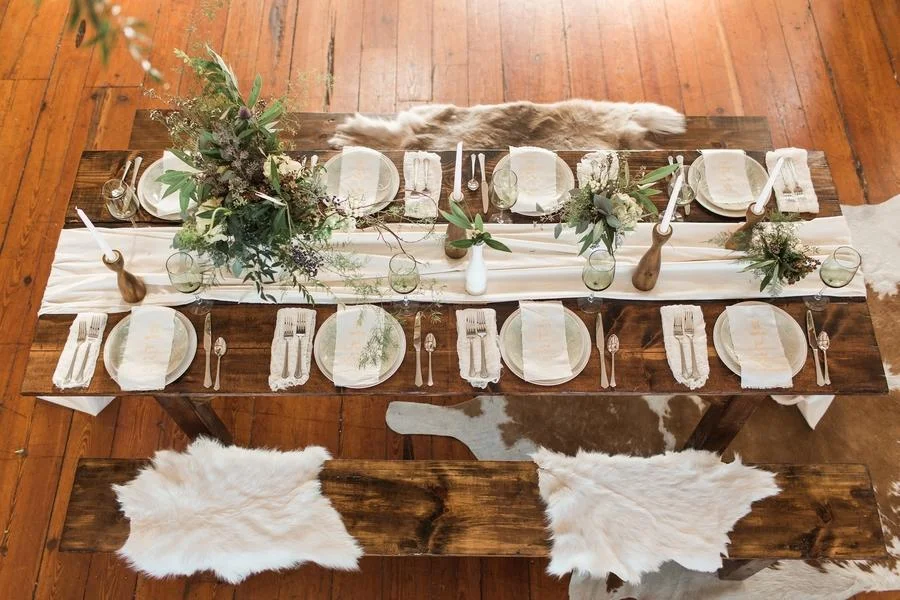 A rustic wooden dining table set for a meal with white plates, silverware, and wine glasses, with a white table runner, decorative vases with green foliage, and tall candles.