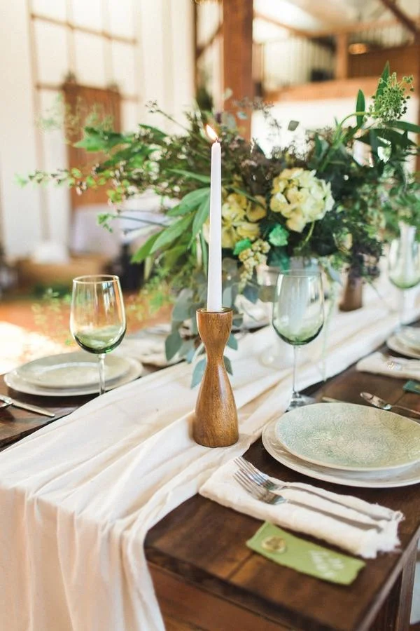 Restaurant table decorated with a white tablecloth, a wooden candle holder with a lit white candle, green wine glasses, ceramic plates, silverware, and a large floral centerpiece with white and green flowers and greenery.