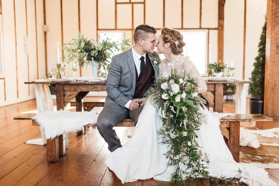 A bride and groom sitting on a wooden bench, kissing, inside a rustic, wooden wedding venue with a decorated table behind them.