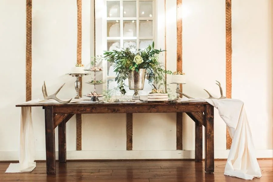 Rustic wooden table decorated for an event with antlers, a large floral arrangement, candles, and draped fabric with a window in the background.