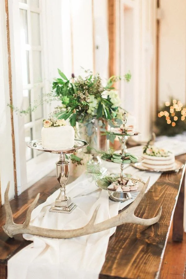Decorative table with cake, macarons, and flowers on a rustic wooden table, with antlers and a white table runner, in a warmly lit room