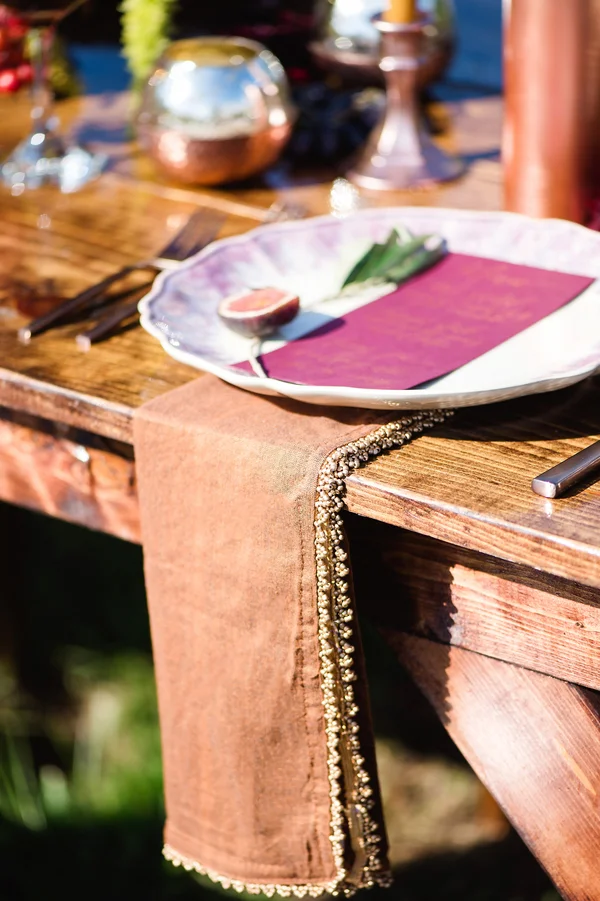 A rustic outdoor table setting with a wooden table pinned with a brown table runner that has an intricate beaded trim, set with a decorative plate, purple napkin, and cutlery.