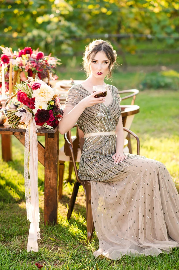 A young woman in an elegant, sparkly gown sits outdoors at a table, holding a glass of red wine, with a backdrop of lush greenery and decorated with colorful flowers.