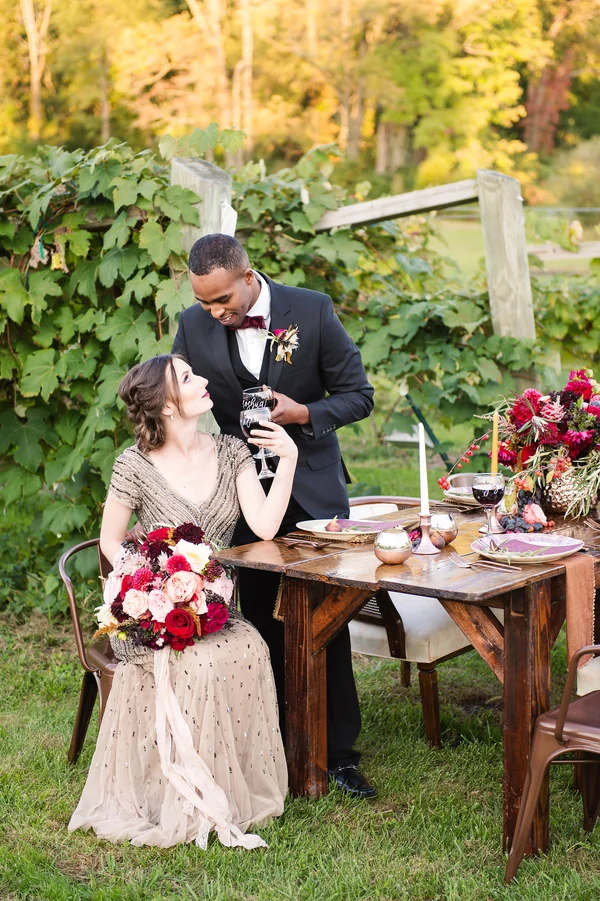 A man in a black tuxedo and a woman in a beige dress sitting at a table outdoors with wine glasses, surrounded by greenery and floral arrangements.