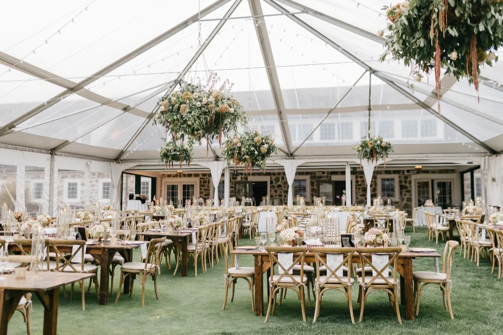 Wide view of a clear-tent wedding reception with long farmhouse tables and floral installations